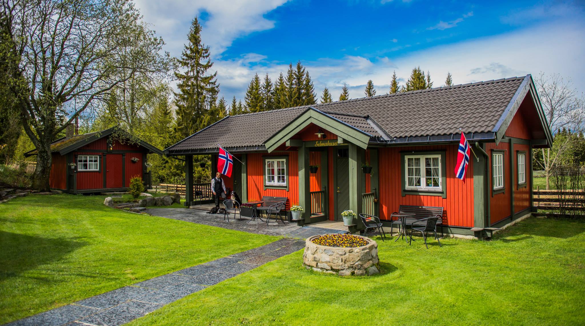 Selmahuset med norgesflagg. En person står med en hund lenger inn i bildet. Bryggehuset kan ses til venstre i bakgrunnen.